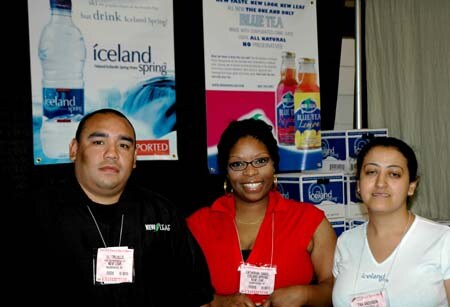 Three people wearing name badges stand at a booth with Iceland Spring water and New Leaf tea posters behind them; the woman in the middle wears a red top and glasses.