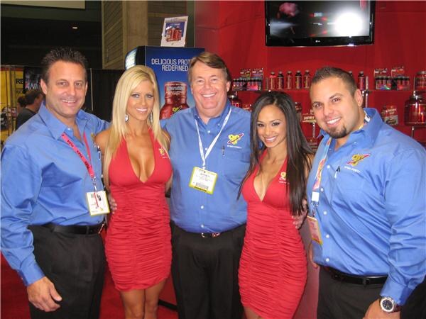 Three men in blue BSN shirts and two women in red dresses stand close together in a row in front of a red display with BSN products on shelves.