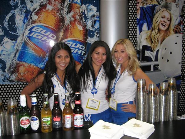 Three smiling women in white tops with lanyards stand behind a bar with Bud Light and Stella Artois bottles, cups and napkins, in front of a large Bud Light bottle graphic.