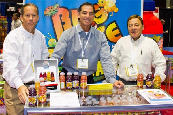 Three men in collared shirts and lanyards stand behind a glass counter of Bug Juice bottles and sample cups, with a colorful Bug Juice banner featuring cartoon bugs behind them.