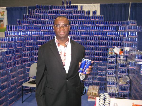 A man in a dark suit and glasses holds two blue Bullet energy drink cans in front of a large wall of stacked Bullet cans.