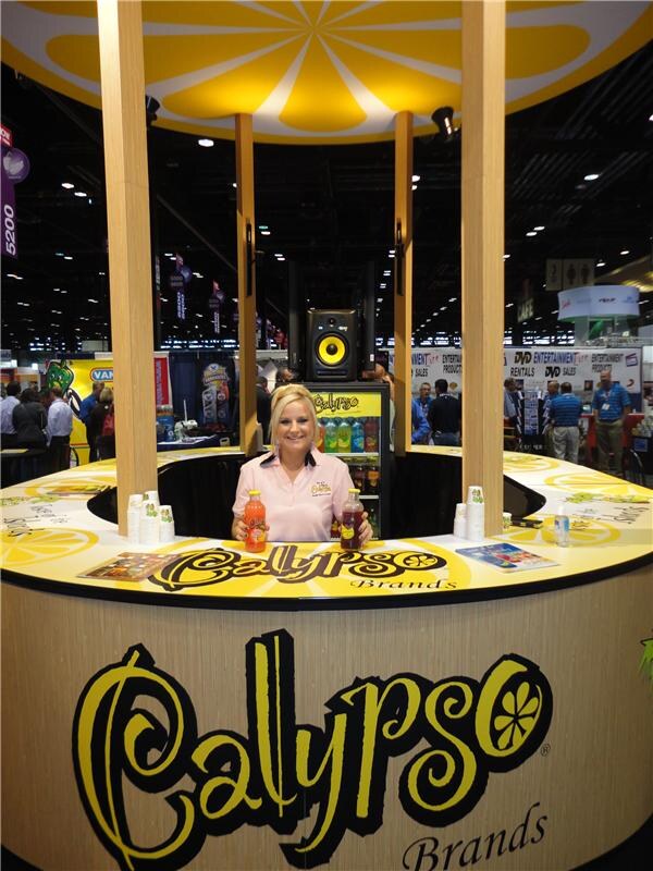 A smiling blonde woman in a pink polo stands inside a round Calypso Brands booth topped with a lemon slice design, holding two bottles of drink. Calypso lettering is on the counter.