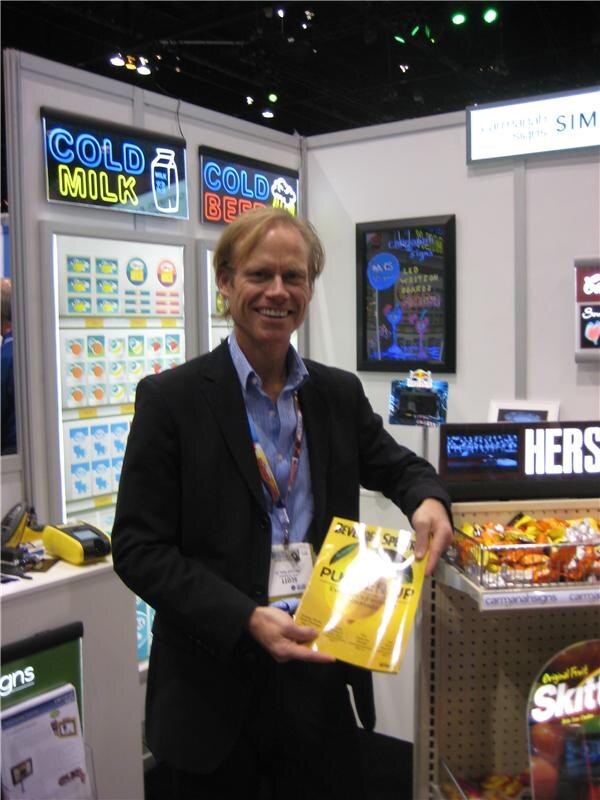 A smiling man in a dark blazer holds a yellow booklet at the Carmanah Signs booth. Illuminated Cold Milk and Cold Beer signs are behind him, with Hershey's and Skittles displays nearby.