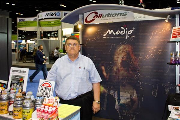 A man in a light blue shirt stands beside a table of supplement bottles at a booth with Cellutions and Modjo signs. A dark Modjo backdrop is behind him.
