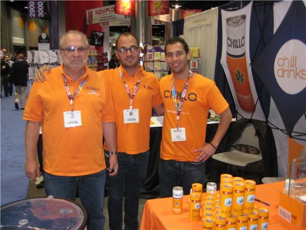 Three men in orange Chillo shirts and badges stand at a booth table topped with Chillo cans. Chill Drinks banners hang behind them.