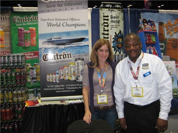 A woman and a man in a white shirt, both wearing badges, pose at the Cintron booth. Banners show a racing boat and stacked Cintron cans.