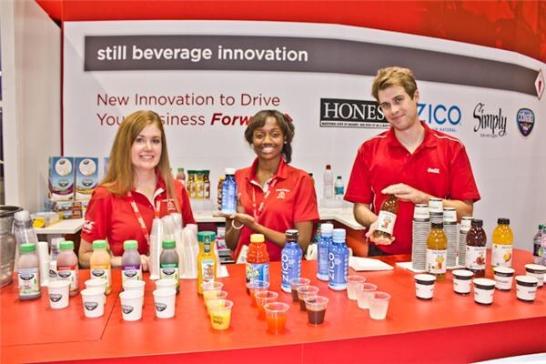 Three people in red shirts stand behind a red counter holding bottles and cups of drinks. Signs behind them show Honest Tea, Zico and Simply under the text still beverage innovation.