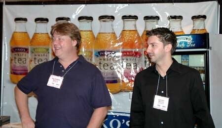 Two men wearing name badges stand in front of a banner showing large Inko's white tea bottles; the man on the left in a dark polo is smiling and looking away.