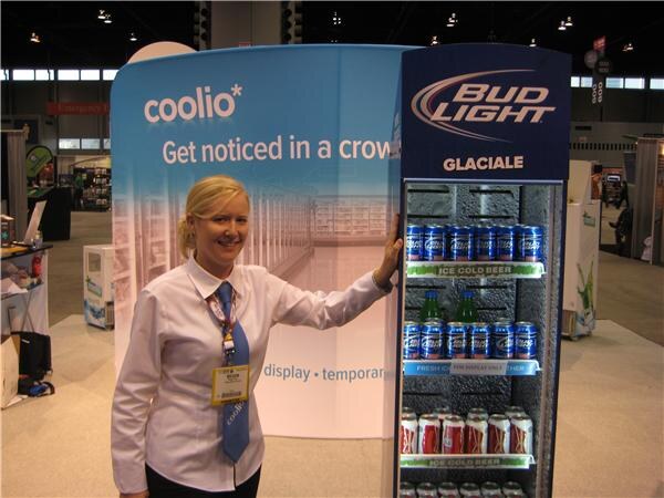 A smiling blonde woman in a white shirt and blue tie leans on a tall Bud Light Glaciale display cooler stocked with cans. A Coolio banner is behind her.