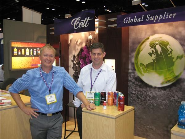 Two men in lanyards stand at a wooden counter holding cans of drink at the Cott booth. A Cott sign, a Global Supplier banner and a globe graphic are behind them.
