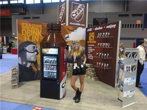 A blonde woman in a cap and dark outfit poses beside a giant Crave bottle cooler at the Crave booth. Banners read It's a Crave new world, drink it in.