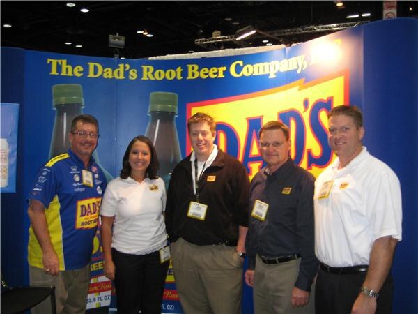 Five people, one woman and four men, pose together in front of a blue Dad's Root Beer Company backdrop showing large bottles and the Dad's logo. They wear name badges.