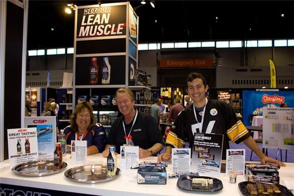 Three people stand behind a white counter at the Detour Lean Muscle booth, with bottles, bars on trays and product signs in front of them. A Stauffer's sign is visible in the background.