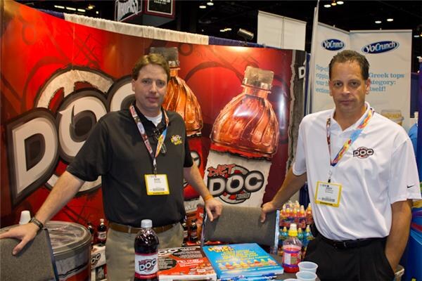 Two men with lanyards and badges stand at a booth with a large red Diet Doc bottle graphic behind them. A bottle of Doc soda sits on the table, and YoCrunch banners hang nearby.