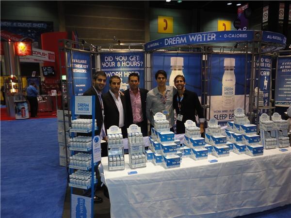 Five men stand behind a draped table covered with Dream Water display boxes. Blue banners above the booth carry the Dream Water name and slogans, and a retail rack stands at left.