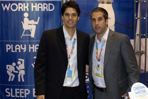 Two men in jackets with convention lanyards and badges stand side by side in front of a blue Dream Water banner reading Work Hard, Play Hard, Sleep Easy. A Dream Water sign is at lower right.