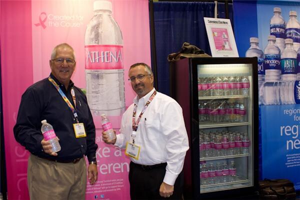Two men with badges hold small pink-labeled Athena water bottles beside a glass-door cooler. Athena and Belmont Springs, Hinckley and Alhambra bottle graphics appear on the booth walls.