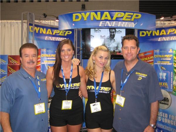 Two men and two women in Dynapep Energy shirts and lanyards pose together under a Dynapep Energy sign. A screen and product packaging are visible behind them.