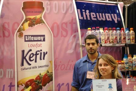 A man and a woman stand at the Lifeway booth beside a large Lowfat Kefir bottle graphic, with shelves of Lifeway bottles behind them.