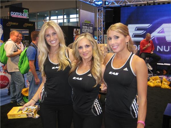 Three smiling women in black EAS tank tops with adidas logos stand in an exhibit hall, one holding a small box, with an EAS screen and other attendees behind them.