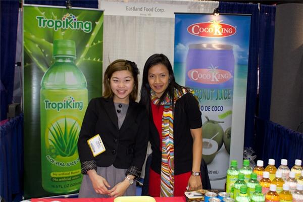 Two women stand at the Eastland Food Corp. booth between a green TropiKing aloe vera banner and a CocoKing coconut juice banner. Colorful bottles are lined up on the table at right.