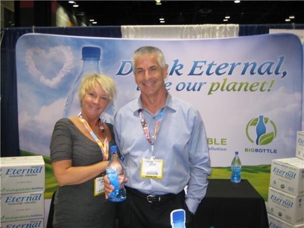 A woman with short blond hair and a man in a blue shirt stand together holding a water bottle in front of a Drink Eternal backdrop with a BioBottle logo and cases of Eternal water.