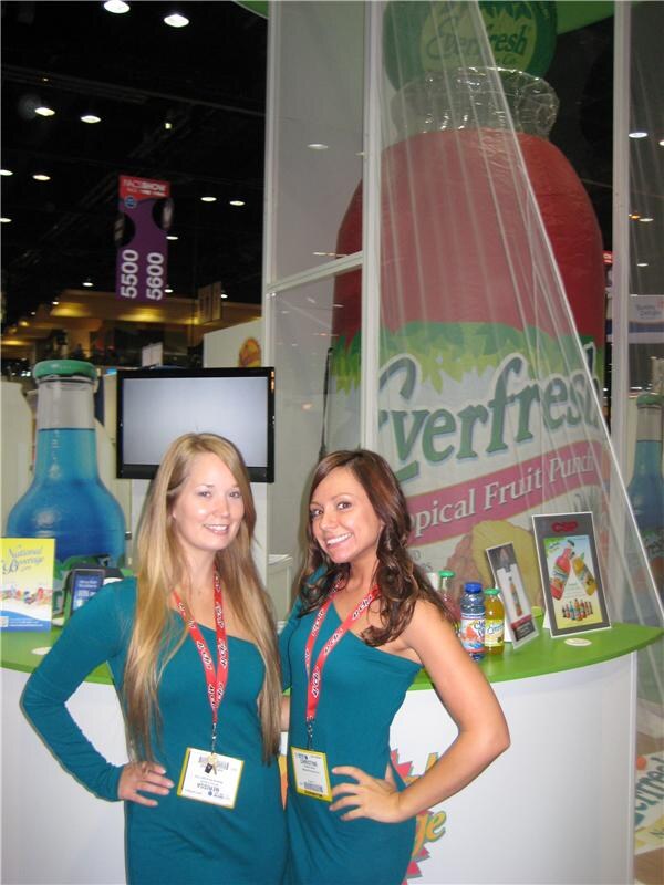 Two smiling women in teal dresses with red lanyards stand at a booth counter in front of a giant inflatable Everfresh Tropical Fruit Punch bottle. A National Beverage sign sits on the counter.