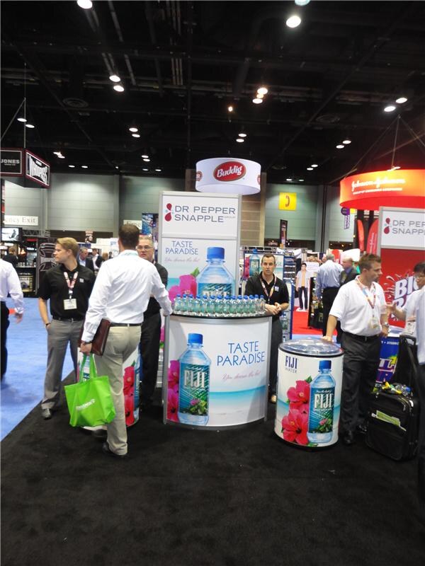 Several men stand around a round Fiji Water display reading Taste Paradise, with Dr Pepper Snapple Group signs, a Buddig hanging sign and a Johnson & Johnson sign overhead.