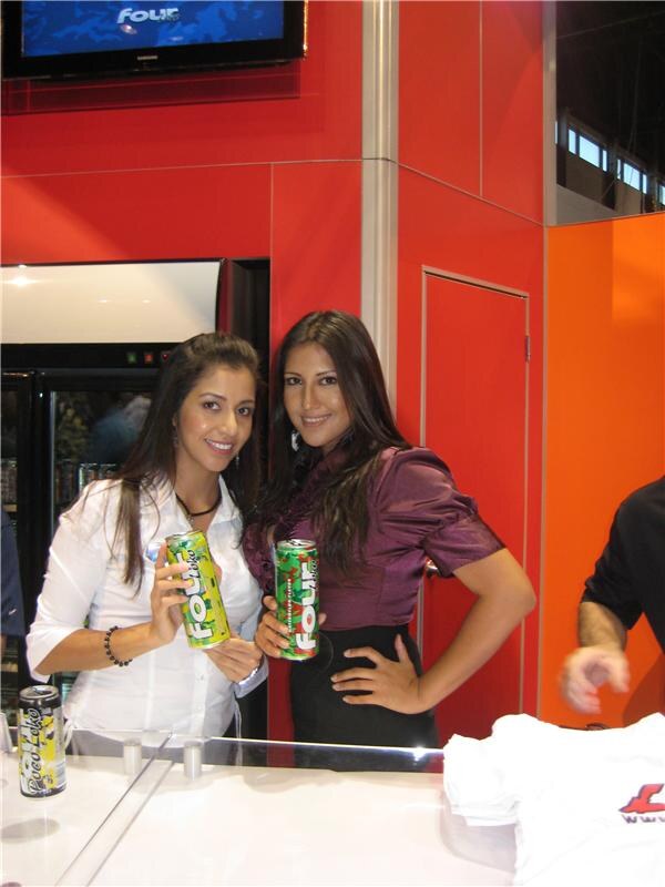 Two women with long dark hair smile and hold cans of Four Loko at a white counter in front of a red wall, with a Four screen above and a white T-shirt on the counter.