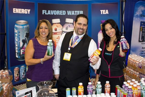 A woman, a man in a vest and a woman in a dark jacket hold bottles of Fresh drinks at a booth with signs for Energy, Flavored Water and Tea, and bottles lined up in front.