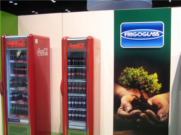 Two red glass-door Coca-Cola coolers stocked with bottles stand beside a green Frigoglass sign showing hands holding a small tree growing from soil.
