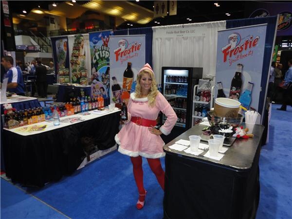 A woman in a pink elf-style costume and red tights holds a bottle at a booth with Frostie banners, tables of bottled sodas and a cooler on a blue carpeted floor.