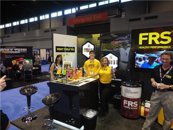 Staff in yellow FRS shirts and a man in a dark FRS polo stand at a display counter of FRS drinks, with a large FRS Healthy Performance sign and a Citrus Pomegranate barrel nearby.