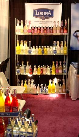 A Lorina sparkling beverages booth with a sign above metal shelves holding rows of bottles in yellow, pink and clear liquids, with more bottles in the foreground.