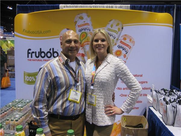 A bald man in a striped shirt and a blond woman in a white textured jacket stand together in front of a Frubob backdrop showing The Natural Fruit Float, with bags and bottles nearby.