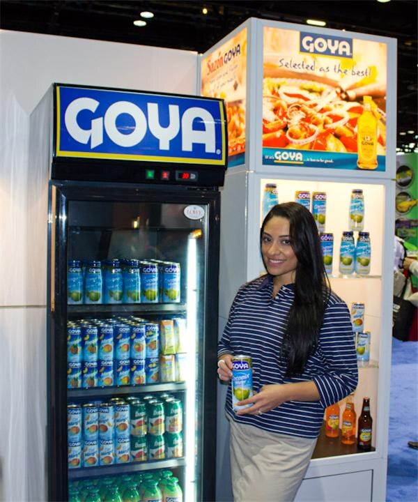 A woman with long dark hair in a striped top holds a can of Goya juice beside a Goya refrigerator and a lit display shelf stocked with Goya cans and bottles.