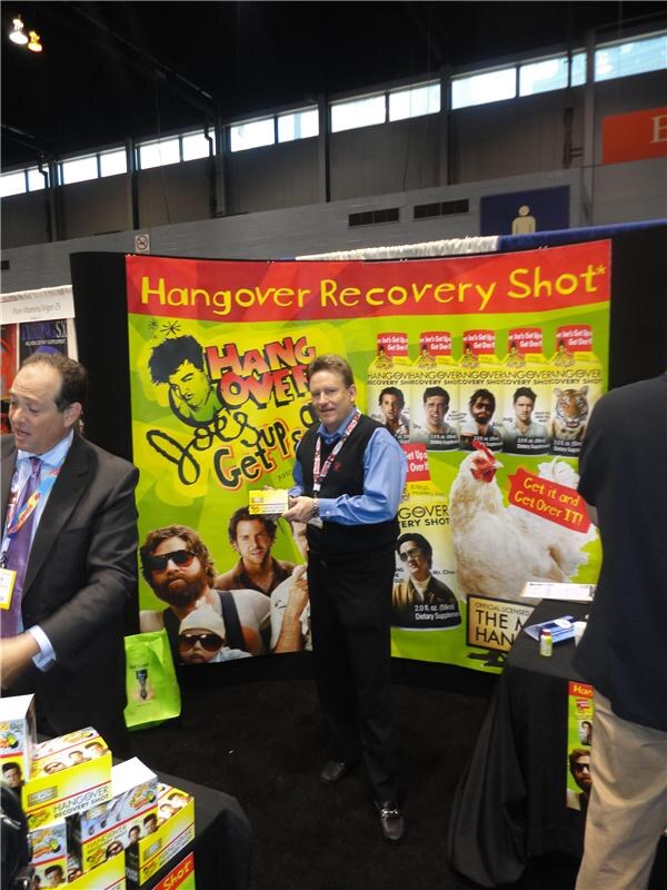 A man in a black vest and blue shirt holds a small box in front of a Hangover Joe's Hangover Recovery Shot banner. A man in a suit stands at left, and product boxes sit on a table.