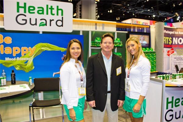 A man in a dark blazer stands between two women in white tops and green shorts in front of HealthGuard signs, including a blue Be Happy Mood Booster banner.