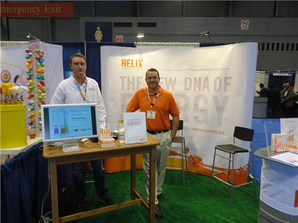 Two men stand beside a wooden table with a monitor in a Helix booth. A banner behind them reads The New DNA of Energy, and the floor is covered in artificial grass.