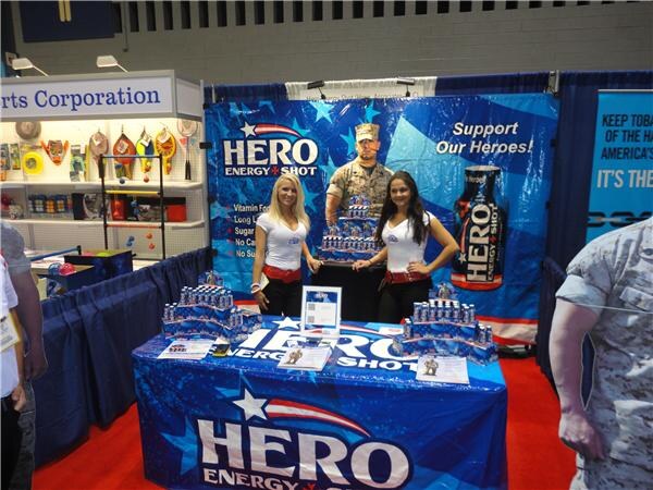 Two women in white shirts stand behind a blue table draped with Hero Energy Shot logos and displays of bottles. A backdrop shows a soldier cutout and the text Support Our Heroes.