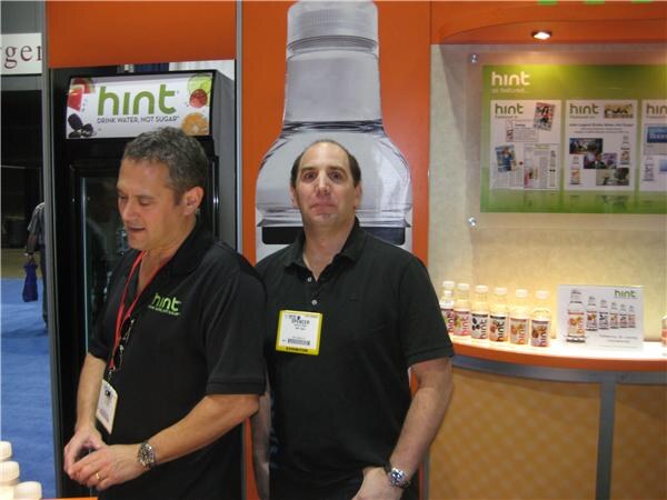 Two men in black polo shirts stand in a Hint booth. One looks down while the other faces the camera. A display of Hint bottles and a cooler are behind them.