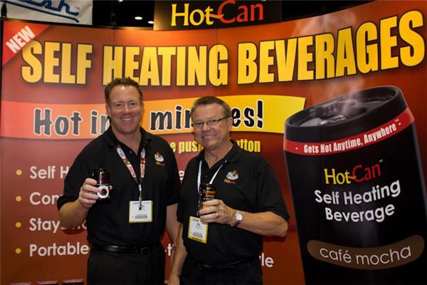 Two smiling men in black polos hold small beverage cans in front of a HotCan banner reading Self Heating Beverages, with a large café mocha can graphic at right.