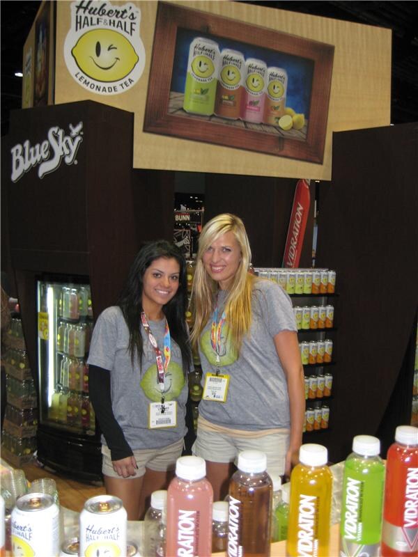 Two women in gray T-shirts stand at a booth with Hubert's Half & Half Lemonade Tea and Blue Sky signs. Vibration bottles and Hubert's cans are on the counter in front.