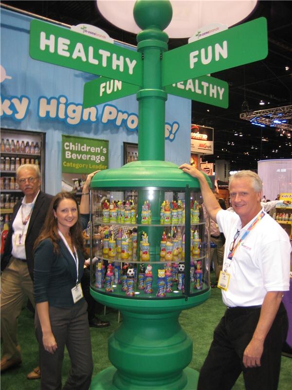 A woman and two men stand around a green signpost-shaped display marked Healthy and Fun, which holds children's beverage bottles. A sign reads Children's Beverage Category Leader.