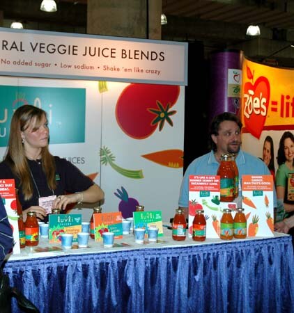 A woman and a man sit behind a table with bottles of veggie juice and small cups under a sign reading veggie juice blends. A Zoe's banner with a red heart is behind them.