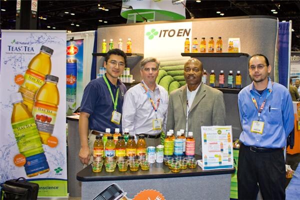 Four men stand behind a counter of bottled teas at the Ito En booth, with a Teas' Tea banner at left and an Ito En sign above shelves of bottles.