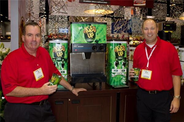 Two men in red polo shirts and badges stand beside Jacked Up ice dispensers with green Jacked Up Energy branding; each holds a green cup.