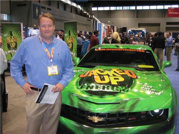 A man in a blue shirt with a badge lanyard stands beside a green Chevrolet Camaro covered in Jacked Up Energy graphics, with Jacked Up banners behind him on the show floor.
