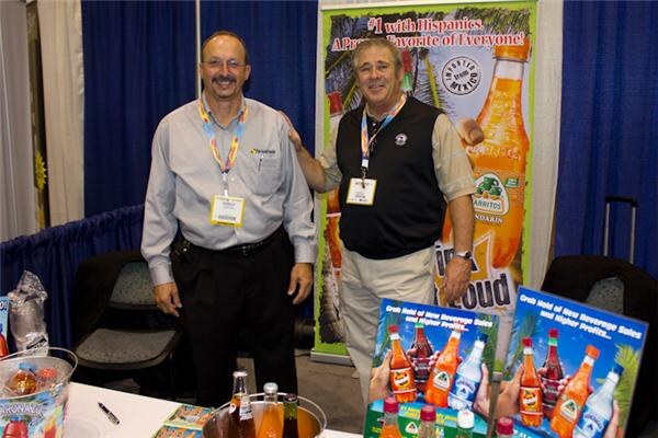 Two smiling men wearing badge lanyards stand at the Jarritos booth, with a Jarritos banner behind them and bottles and promotional signs on the table in front.