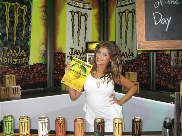 A smiling woman in a white dress holds a Beverage Spectrum magazine in front of Java Monster banners, with a row of Java Monster cans on the counter below.